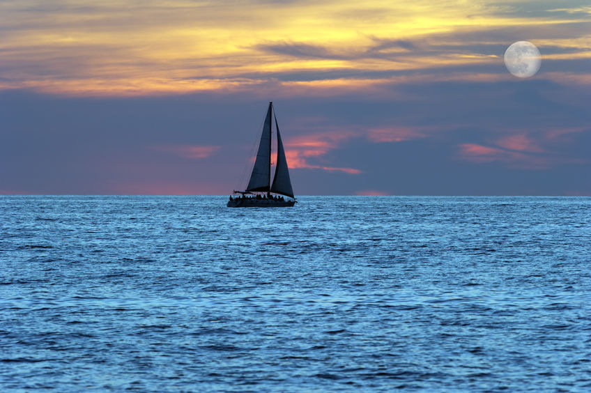 44786395 - sailboat silhouette is a sailboat moving along the water at sunset with a full moon rising in a cloud filled sky.